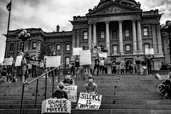 A group of people are gathered on the steps of a large, classical-style building, engaged in a protest. Many are holding signs with messages advocating for justice and equality, such as 'Black Lives Matter', 'No Justice No Peace', and 'End Police Brutality'. The participants appear to be united in their cause, standing on and around the steps with a strong sense of purpose.