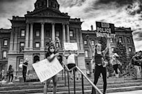 A group of people stands in front of a large government building, holding signs advocating for racial equality and an end to police brutality. The scene is lively with people engaged in peaceful protest. The building in the background features grand columns and intricate architectural details.