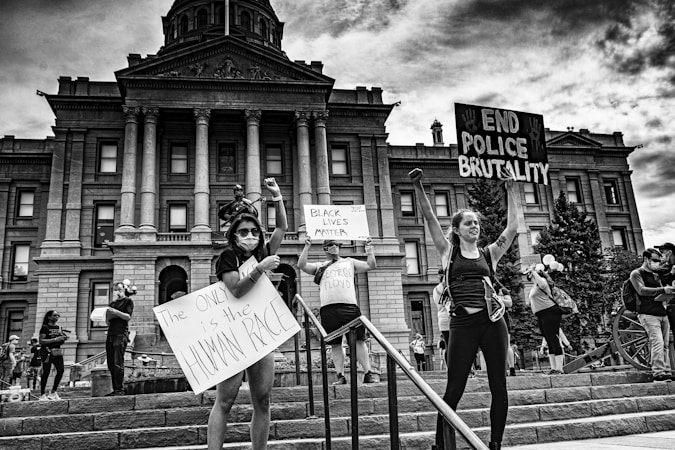 A group of people stands in front of a large government building, holding signs advocating for racial equality and an end to police brutality. The scene is lively with people engaged in peaceful protest. The building in the background features grand columns and intricate architectural details.