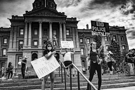A group of people stands in front of a large government building, holding signs advocating for racial equality and an end to police brutality. The scene is lively with people engaged in peaceful protest. The building in the background features grand columns and intricate architectural details.