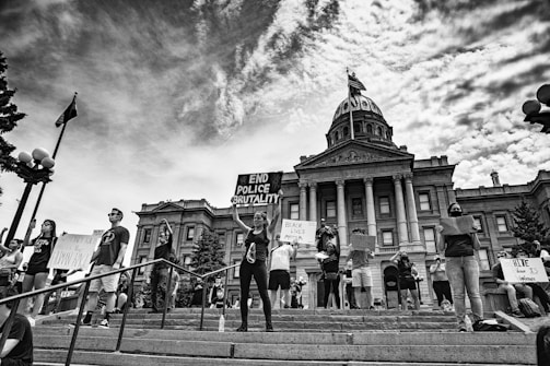 A group of protesters stand on the steps in front of a historic government building holding signs with messages advocating against police brutality and racial injustice. The scene is black and white, capturing a strong sense of activism and unity among the individuals. The architecture of the building includes large columns and a central dome, indicating significance and authority.