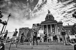 A group of protesters stand on the steps in front of a historic government building holding signs with messages advocating against police brutality and racial injustice. The scene is black and white, capturing a strong sense of activism and unity among the individuals. The architecture of the building includes large columns and a central dome, indicating significance and authority.