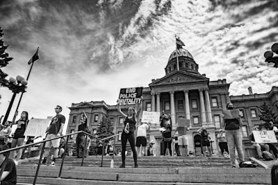 A group of protesters stand on the steps in front of a historic government building holding signs with messages advocating against police brutality and racial injustice. The scene is black and white, capturing a strong sense of activism and unity among the individuals. The architecture of the building includes large columns and a central dome, indicating significance and authority.