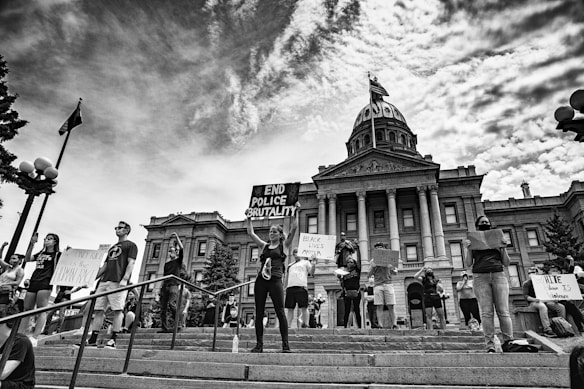 A group of protesters stand on the steps in front of a historic government building holding signs with messages advocating against police brutality and racial injustice. The scene is black and white, capturing a strong sense of activism and unity among the individuals. The architecture of the building includes large columns and a central dome, indicating significance and authority.