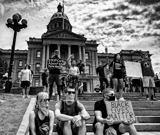 A group of people stands in front of a large government building, holding signs with messages advocating for racial equality. The atmosphere is charged, with participants wearing masks and raising their fists. The scene appears to be a protest or demonstration.
