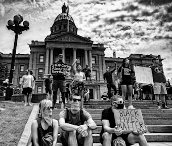 A group of people stands in front of a large government building, holding signs with messages advocating for racial equality. The atmosphere is charged, with participants wearing masks and raising their fists. The scene appears to be a protest or demonstration.