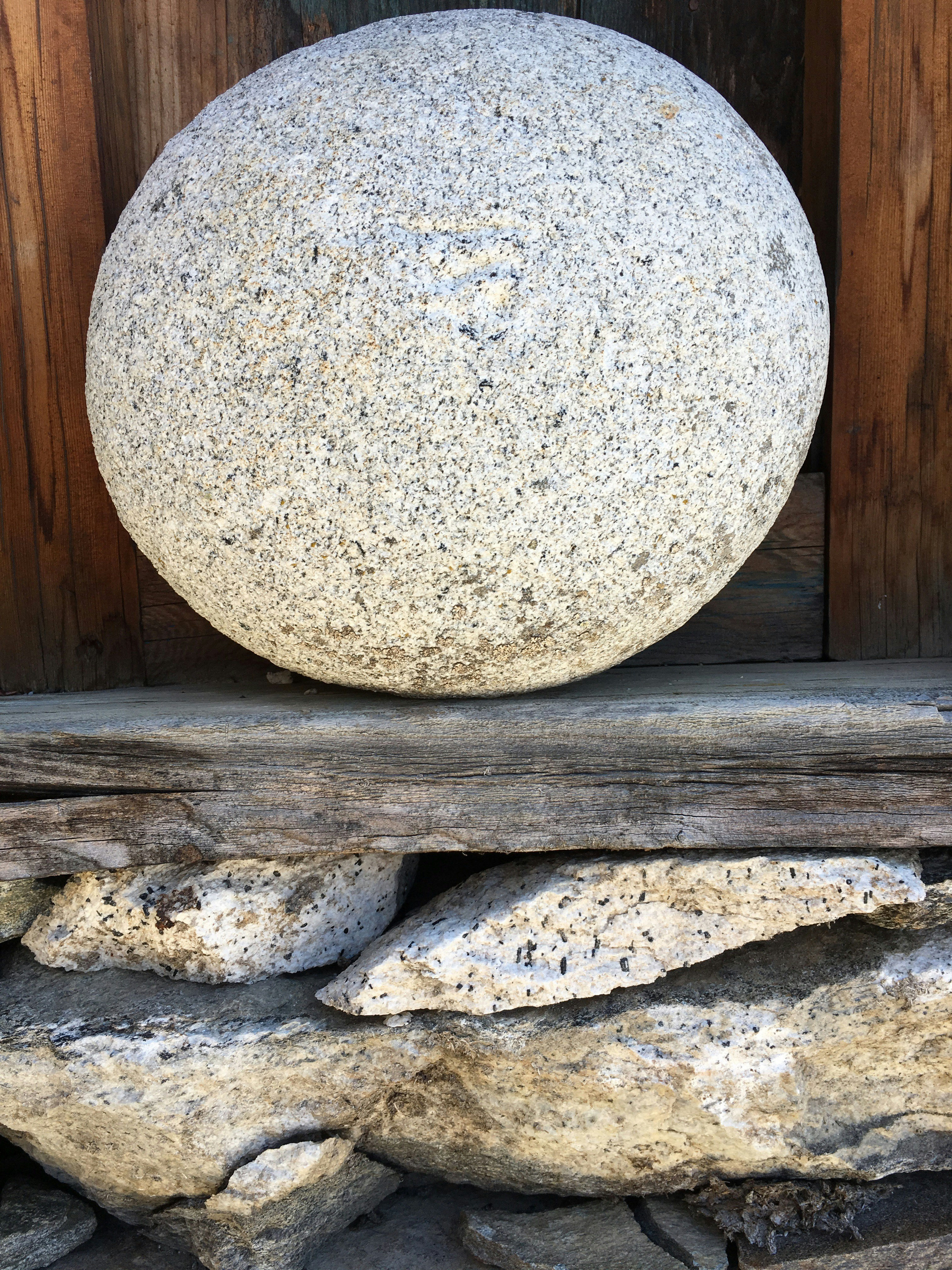 A large, spherical stone with a textured surface rests on a stack of wooden and stone supports. The stone appears weathered, with a natural gray and white speckled pattern. The background includes dark wood panels, contributing to a rustic and earthy atmosphere.