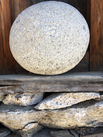 A large, spherical stone with a textured surface rests on a stack of wooden and stone supports. The stone appears weathered, with a natural gray and white speckled pattern. The background includes dark wood panels, contributing to a rustic and earthy atmosphere.