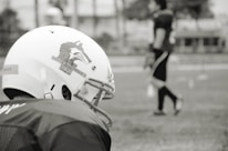 A football player is seen wearing a helmet with a wolf logo and the text 'Wolves Peru' on the side. In the background, another player stands blurred on a grassy field, suggesting a practice or game setting.