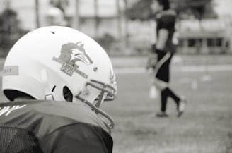 A football player is seen wearing a helmet with a wolf logo and the text 'Wolves Peru' on the side. In the background, another player stands blurred on a grassy field, suggesting a practice or game setting.