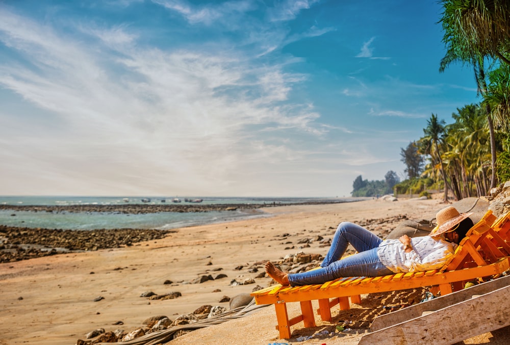 woman in white shirt and blue denim jeans lying on orange bench on beach shore during