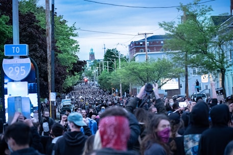 A large crowd of people fills a city street during a demonstration. Many individuals are holding up signs and taking pictures or videos with their phones. The street is flanked by trees and buildings, and there are power lines above. The atmosphere is energetic and engaged.