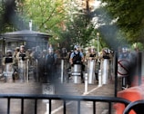 A group of law enforcement officers in riot gear, including helmets and shields, stand behind a barrier in a street setting with trees in the background. There are visible signs indicating no bicycles and military presence.