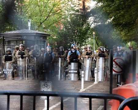 A group of law enforcement officers in riot gear, including helmets and shields, stand behind a barrier in a street setting with trees in the background. There are visible signs indicating no bicycles and military presence.