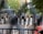 A group of law enforcement officers in riot gear, including helmets and shields, stand behind a barrier in a street setting with trees in the background. There are visible signs indicating no bicycles and military presence.