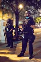 Two individuals dressed in full police riot gear with helmets and face shields stand on a city sidewalk at night. They appear vigilant, holding batons and wearing black uniforms. The background includes a tree, some buildings, and streetlights illuminating the scene.