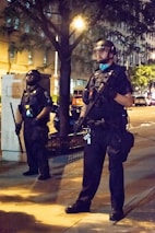Two individuals dressed in full police riot gear with helmets and face shields stand on a city sidewalk at night. They appear vigilant, holding batons and wearing black uniforms. The background includes a tree, some buildings, and streetlights illuminating the scene.