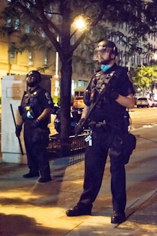 Two individuals dressed in full police riot gear with helmets and face shields stand on a city sidewalk at night. They appear vigilant, holding batons and wearing black uniforms. The background includes a tree, some buildings, and streetlights illuminating the scene.