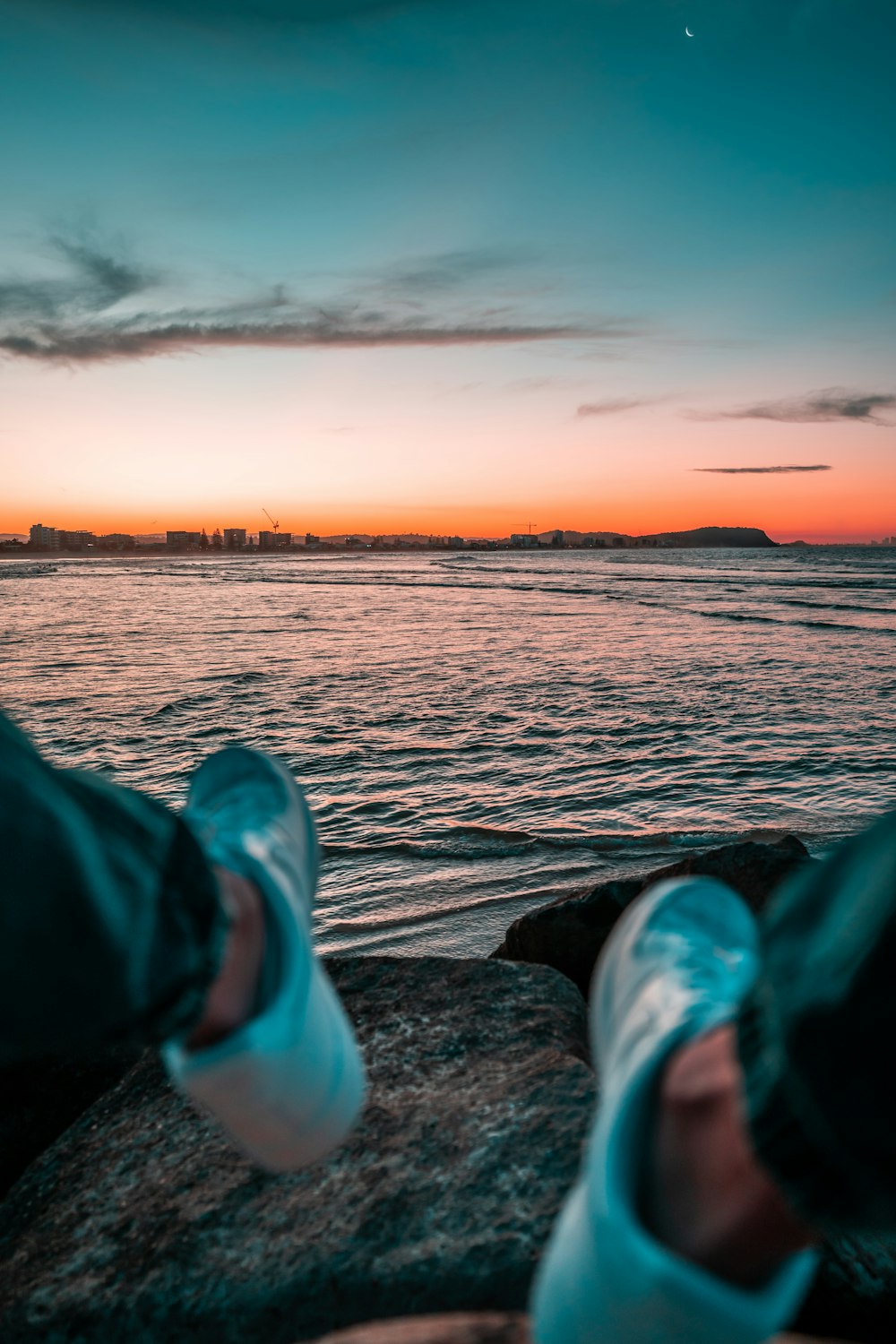 person in green pants and white sneakers sitting on rock near body of water during sunset