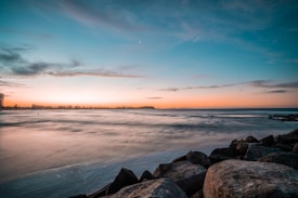 A tranquil coastline with a rocky foreground, gently lapping waves, and a sprawling city skyline in the distance under a gradient sky transitioning from deep blue to warm orange tones.