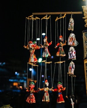 A collection of colorful puppets hangs from strings in a dimly lit setting. The puppets are dressed in bright traditional costumes with intricate designs. The background is blurry, with soft circular lights that create a festive atmosphere.