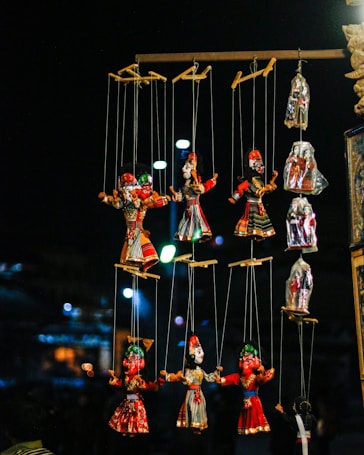 A collection of colorful puppets hangs from strings in a dimly lit setting. The puppets are dressed in bright traditional costumes with intricate designs. The background is blurry, with soft circular lights that create a festive atmosphere.