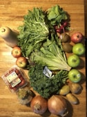 A collection of fresh fruits and vegetables is arranged on a wooden table. The assortment includes a butternut squash, a variety of greens like kale and lettuce, red radishes, apples, kiwis, onions, potatoes, and a container of organic grape tomatoes. The vibrant colors and textures are highlighted by natural light.