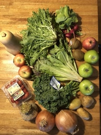 A collection of fresh fruits and vegetables is arranged on a wooden table. The assortment includes a butternut squash, a variety of greens like kale and lettuce, red radishes, apples, kiwis, onions, potatoes, and a container of organic grape tomatoes. The vibrant colors and textures are highlighted by natural light.