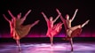 Four dancers are performing in synchrony on a stage with a dark backdrop and a pinkish-purple glow. Each dancer is dressed in flowing, earth-toned costumes, and they are captured in mid-movement with one leg extended high and arms gracefully outstretched. The lighting casts soft shadows on the floor, adding depth and highlighting their elegant poses.