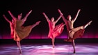 Four dancers are performing in synchrony on a stage with a dark backdrop and a pinkish-purple glow. Each dancer is dressed in flowing, earth-toned costumes, and they are captured in mid-movement with one leg extended high and arms gracefully outstretched. The lighting casts soft shadows on the floor, adding depth and highlighting their elegant poses.