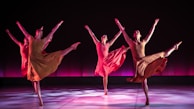 Four dancers are performing in synchrony on a stage with a dark backdrop and a pinkish-purple glow. Each dancer is dressed in flowing, earth-toned costumes, and they are captured in mid-movement with one leg extended high and arms gracefully outstretched. The lighting casts soft shadows on the floor, adding depth and highlighting their elegant poses.