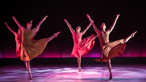 Four dancers are performing in synchrony on a stage with a dark backdrop and a pinkish-purple glow. Each dancer is dressed in flowing, earth-toned costumes, and they are captured in mid-movement with one leg extended high and arms gracefully outstretched. The lighting casts soft shadows on the floor, adding depth and highlighting their elegant poses.