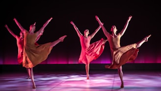 Four dancers are performing in synchrony on a stage with a dark backdrop and a pinkish-purple glow. Each dancer is dressed in flowing, earth-toned costumes, and they are captured in mid-movement with one leg extended high and arms gracefully outstretched. The lighting casts soft shadows on the floor, adding depth and highlighting their elegant poses.