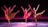 Four dancers are performing in synchrony on a stage with a dark backdrop and a pinkish-purple glow. Each dancer is dressed in flowing, earth-toned costumes, and they are captured in mid-movement with one leg extended high and arms gracefully outstretched. The lighting casts soft shadows on the floor, adding depth and highlighting their elegant poses.