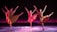 Four dancers are performing in synchrony on a stage with a dark backdrop and a pinkish-purple glow. Each dancer is dressed in flowing, earth-toned costumes, and they are captured in mid-movement with one leg extended high and arms gracefully outstretched. The lighting casts soft shadows on the floor, adding depth and highlighting their elegant poses.
