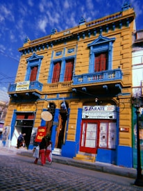A brightly colored yellow and blue building with red windows stands prominently along a cobblestone street. The architecture features ornate details and multiple balconies. Signs indicate a restaurant and a wax museum, with people walking past carrying shopping bags.