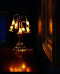 A delicately crafted lamp with multiple bell-shaped glass shades emits a warm, golden glow. The lamp sits on a polished wooden surface, and the rich, dark background adds contrast to the ambient light. The reflection of the lamp's light creates a soft, inviting atmosphere.