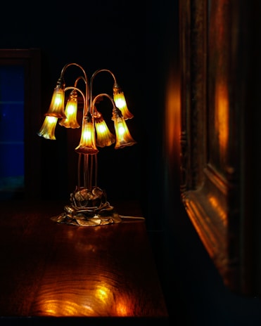 A delicately crafted lamp with multiple bell-shaped glass shades emits a warm, golden glow. The lamp sits on a polished wooden surface, and the rich, dark background adds contrast to the ambient light. The reflection of the lamp's light creates a soft, inviting atmosphere.