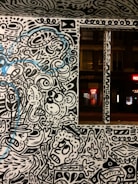 A wall adorned with vibrant street art featuring abstract and whimsical characters and patterns. The artwork is primarily in black and white with some blue accents. The scene includes stylized faces, swirling lines, and various abstract forms, creating a lively and dynamic visual experience.