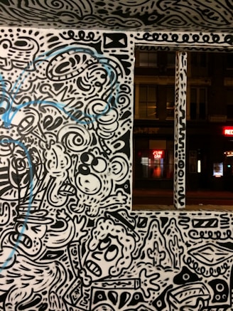 A wall adorned with vibrant street art featuring abstract and whimsical characters and patterns. The artwork is primarily in black and white with some blue accents. The scene includes stylized faces, swirling lines, and various abstract forms, creating a lively and dynamic visual experience.