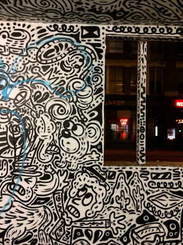 A wall adorned with vibrant street art featuring abstract and whimsical characters and patterns. The artwork is primarily in black and white with some blue accents. The scene includes stylized faces, swirling lines, and various abstract forms, creating a lively and dynamic visual experience.
