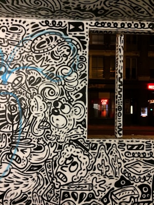 A wall adorned with vibrant street art featuring abstract and whimsical characters and patterns. The artwork is primarily in black and white with some blue accents. The scene includes stylized faces, swirling lines, and various abstract forms, creating a lively and dynamic visual experience.