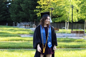 A young person is standing in a grassy outdoor setting, wearing a graduation cap and gown with a blue stole and gold tassels. The background features lush, green trees and a wooden fence.