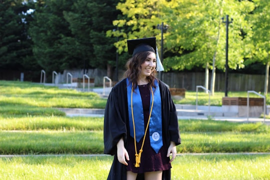 A young person is standing in a grassy outdoor setting, wearing a graduation cap and gown with a blue stole and gold tassels. The background features lush, green trees and a wooden fence.