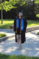 A person is wearing a graduation cap and gown, accompanied by a blue stole and gold cords. They are standing on a paved pathway surrounded by lush green grass and trees, with sunlight filtering through the leaves, casting shadows on the ground.