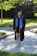 A person is wearing a graduation cap and gown, accompanied by a blue stole and gold cords. They are standing on a paved pathway surrounded by lush green grass and trees, with sunlight filtering through the leaves, casting shadows on the ground.