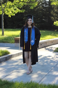 A person is wearing a graduation cap and gown, accompanied by a blue stole and gold cords. They are standing on a paved pathway surrounded by lush green grass and trees, with sunlight filtering through the leaves, casting shadows on the ground.