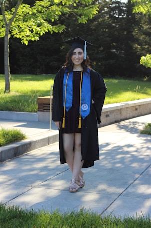 A person is wearing a graduation cap and gown, accompanied by a blue stole and gold cords. They are standing on a paved pathway surrounded by lush green grass and trees, with sunlight filtering through the leaves, casting shadows on the ground.