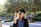 A graduate in cap and gown holds a sign with '#DONE' while standing outdoors. The background features lush greenery and appears to be on a sunny day.