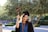 A graduate in cap and gown holds a sign with '#DONE' while standing outdoors. The background features lush greenery and appears to be on a sunny day.
